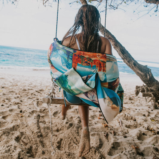A girl sits on a wooden beach swing with a colorful printed towel around her, with sand, ocean, and a tree in the background.