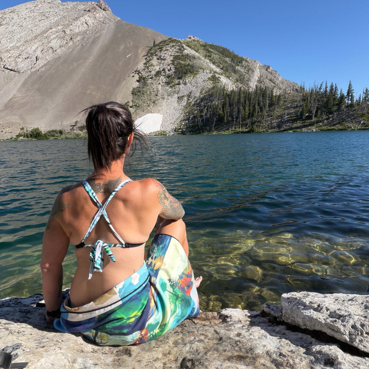 A woman sits on a rocky lakeshore with her back to the camera, wearing a multicolor print towel and a black bikini with light blue straps, looking out over a mountain and blue lake.