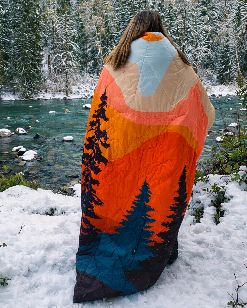 A woman stands in a snowy outdoor landscape with a river and evergreen forest in the background, wrapped in a colorful blanket featuring an orange, blue, black, and beige mountain-and-trees design.
