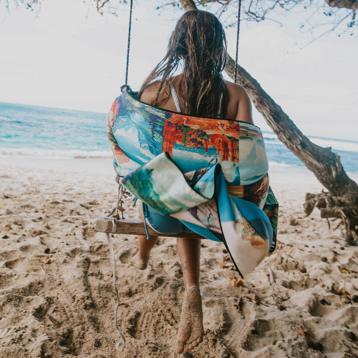 A girl sits on a wooden beach swing with a colorful printed towel around her, with sand, ocean, and a tree in the background.