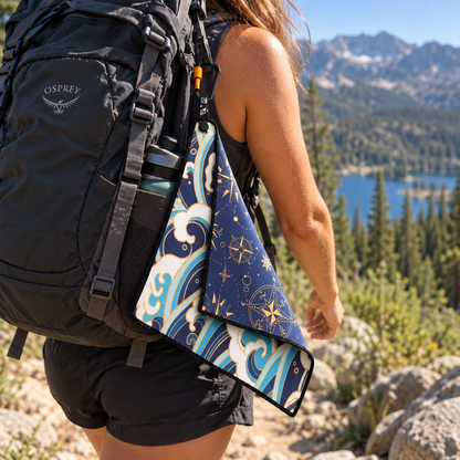 A person hiking outdoors on a rocky mountain trail wearing an Osprey backpack and carrying a DryFoxCo washcloth with a waves and compass print. A lake and forest are visible in the background.