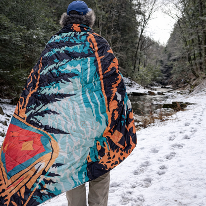 A person standing in a snowy outdoor setting near a stream, wrapped in a large quilted blanket with a bold abstract pattern in teal, orange, black, and red.