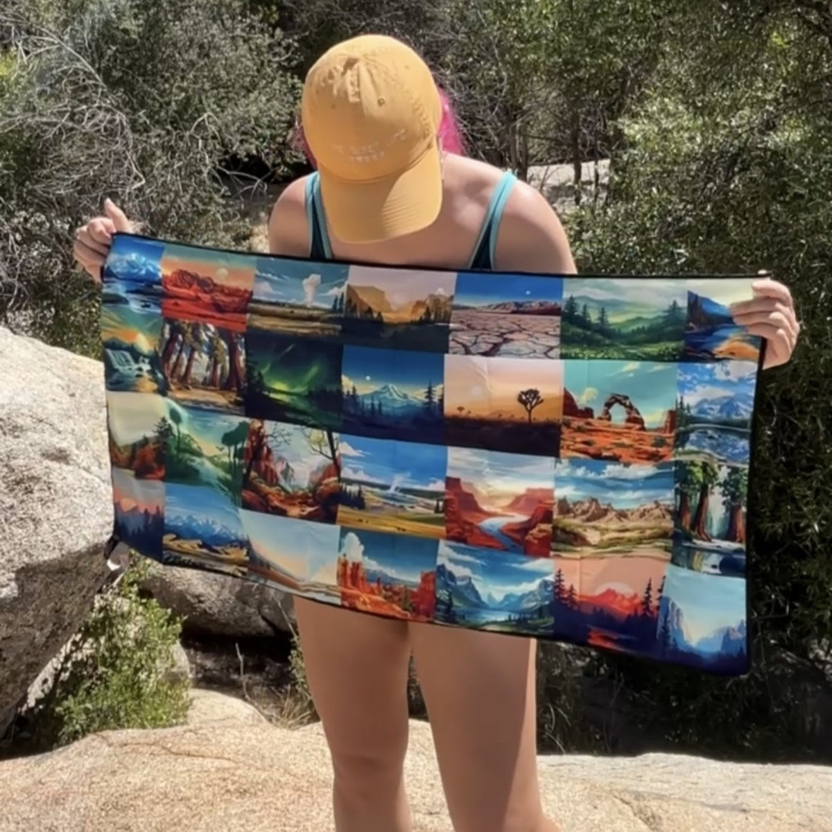 A person outdoors holding up a DryFox Pack towel in Explore print with a grid of colorful landscape photos, including red rock canyons, mountains, lakes, and desert scenes.