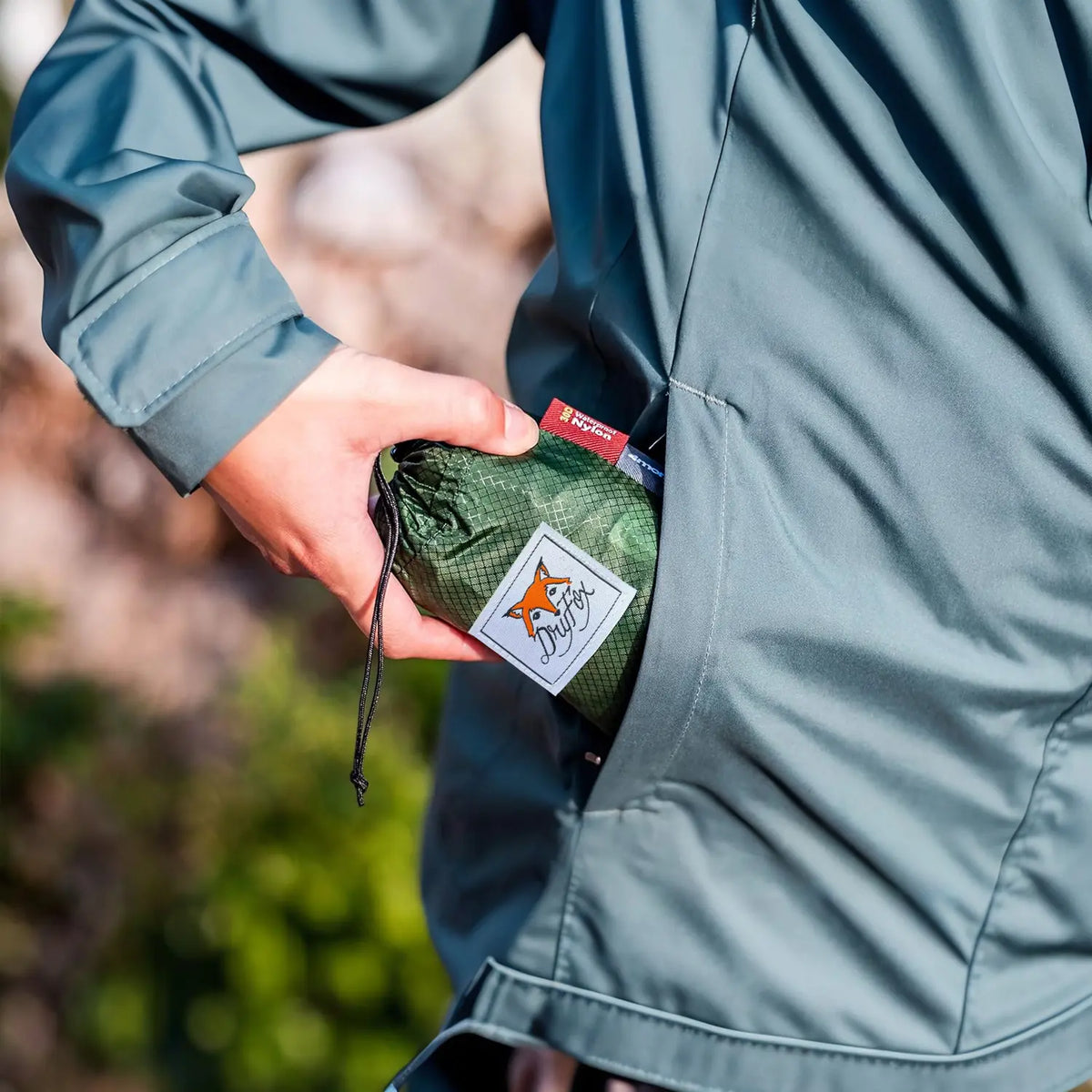 A close-up lifestyle shot of a person wearing a slate blue-gray jacket and holding a small green mesh drawstring pouch with a white Fox Pouch label. The person is shown from the side, with a blurred outdoor background.