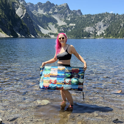 A woman with bright pink hair stands in shallow water at a mountain lake, holding up a DryFox Pack towel in Explore print with colorful scenic prints of the US national parks.