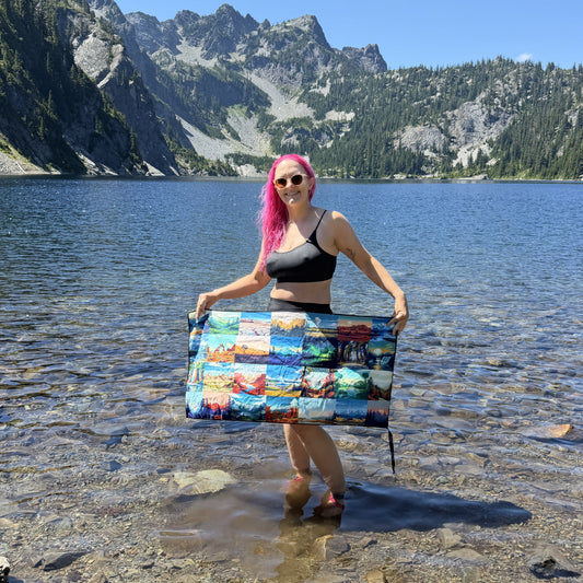 A woman with bright pink hair stands in shallow water at a mountain lake, holding up a DryFox Pack towel in Explore print with colorful scenic prints of the US national parks.
