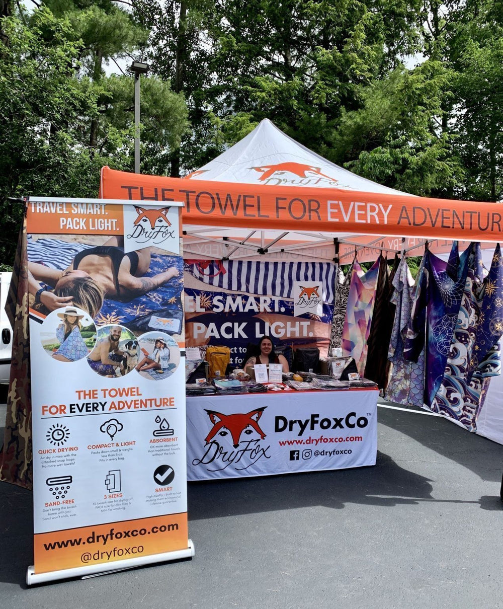 Outdoor market booth for DryFoxCo featuring branded tents, roll-up display signs, and hanging towels on a paved area with trees in the background.