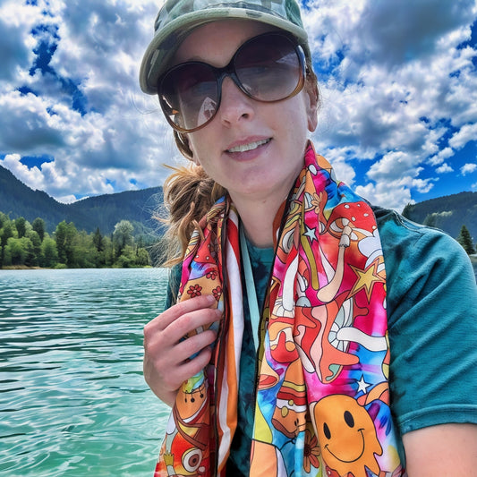 A woman outdoors by a lake wearing a colorful DryFox Pack towel in Trippy print around her neck, with mountains and a cloudy blue sky in the background.