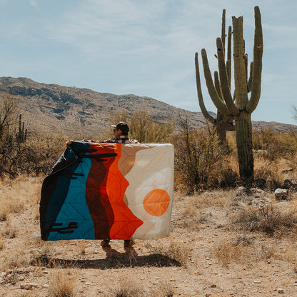 A man stands in a desert landscape holding up a large, folded or draped textile with a bold abstract design in blue, brown, orange, and white. A tall saguaro cactus and mountains are visible in the background under a blue sky.