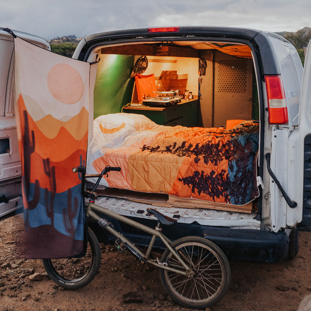 A white van with its rear doors open, styled as a mobile bedroom with a bed inside, warm ambient lighting, and a colorful desert landscape print draped over the side door. A bicycle is parked in front on dirt ground under an overcast sky.