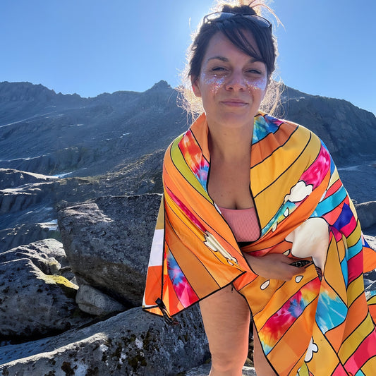 A woman outdoors on rocky mountains wrapped in a bright multicolor patterned towel with orange, yellow, pink, blue, and white abstract shapes.