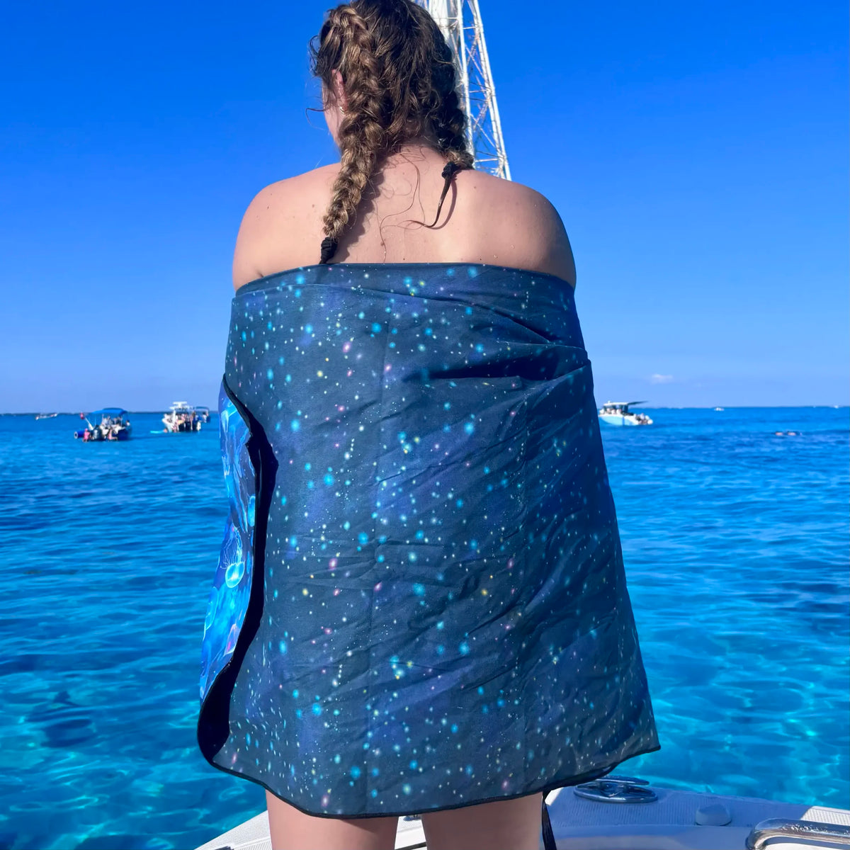 A woman on a boat seen from behind, wrapped in a dark blue towel with luminescent print, overlooking bright turquoise ocean water under a clear blue sky.