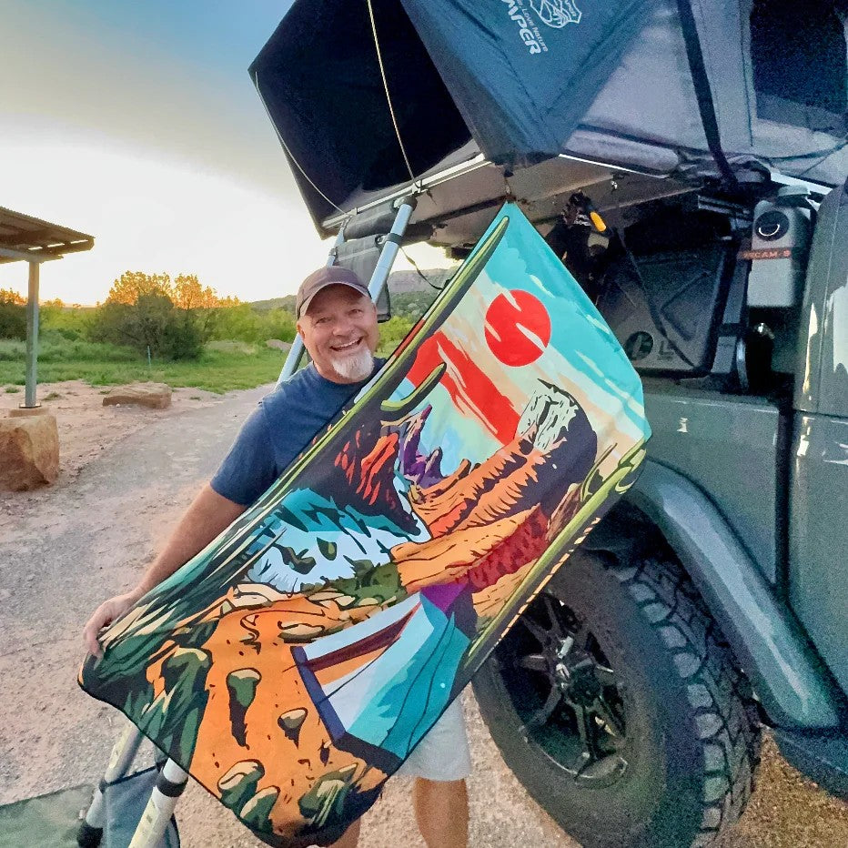 A man outdoors in front of a pop-up rooftop car tent, holding colorful DryFox towel in Campout print illustrated with desert landscape design and a red sun graphic.
